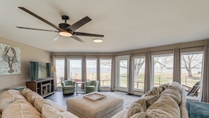 A spacious living room with a large sectional sofa, a coffee table, and a ceiling fan. There are multiple windows providing a view of the outdoors. - Private Beach & Fishing Pier: Wicomico River House (Princess Anne)