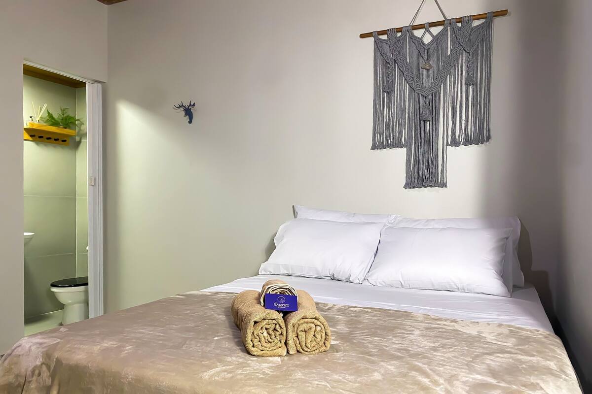 Un lit bien fait, avec des draps blancs, une décoration murale en macramé et une serviette enroulée posée sur le lit.