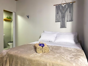A neatly made bed with white linens, a macrame wall hanging, and a rolled towel on the bed. - Pousada Quartzo (Alto Paraíso de Goiás)