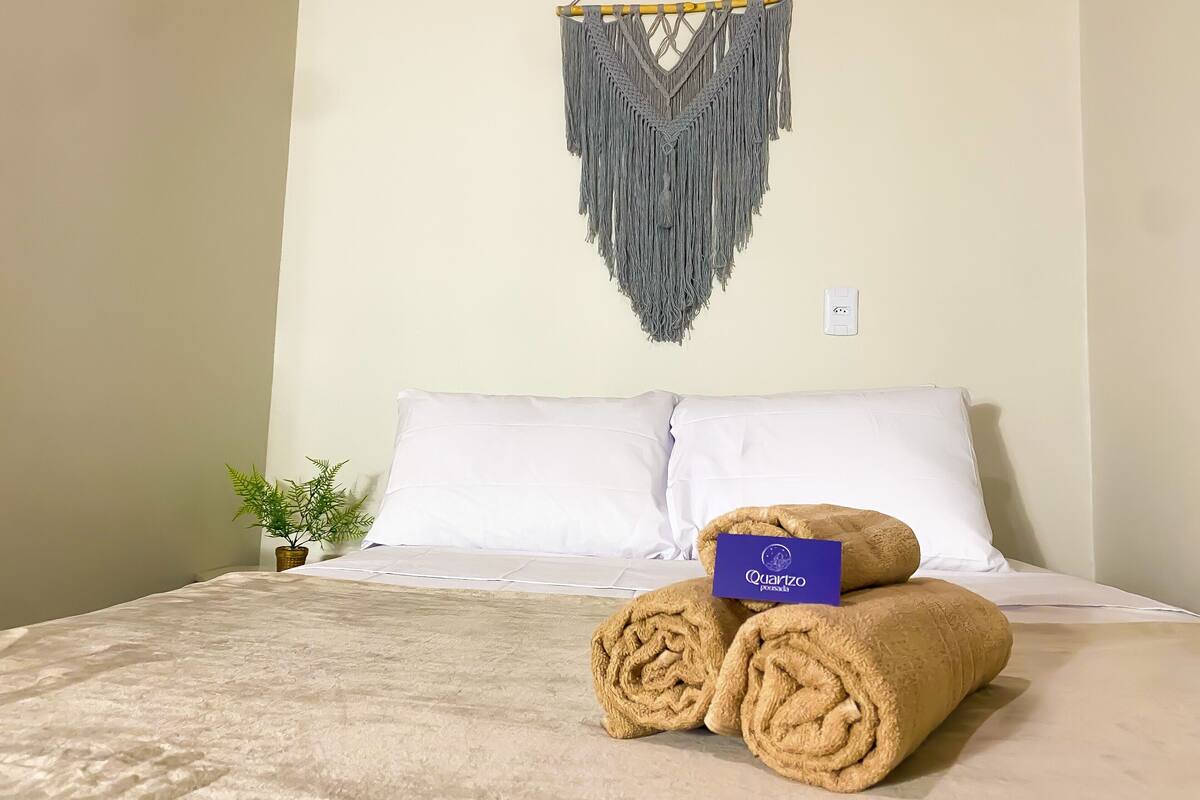 Un lit bien fait, avec des serviettes pliées et une étiquette, une décoration murale en macramé et une petite plante sur le côté.