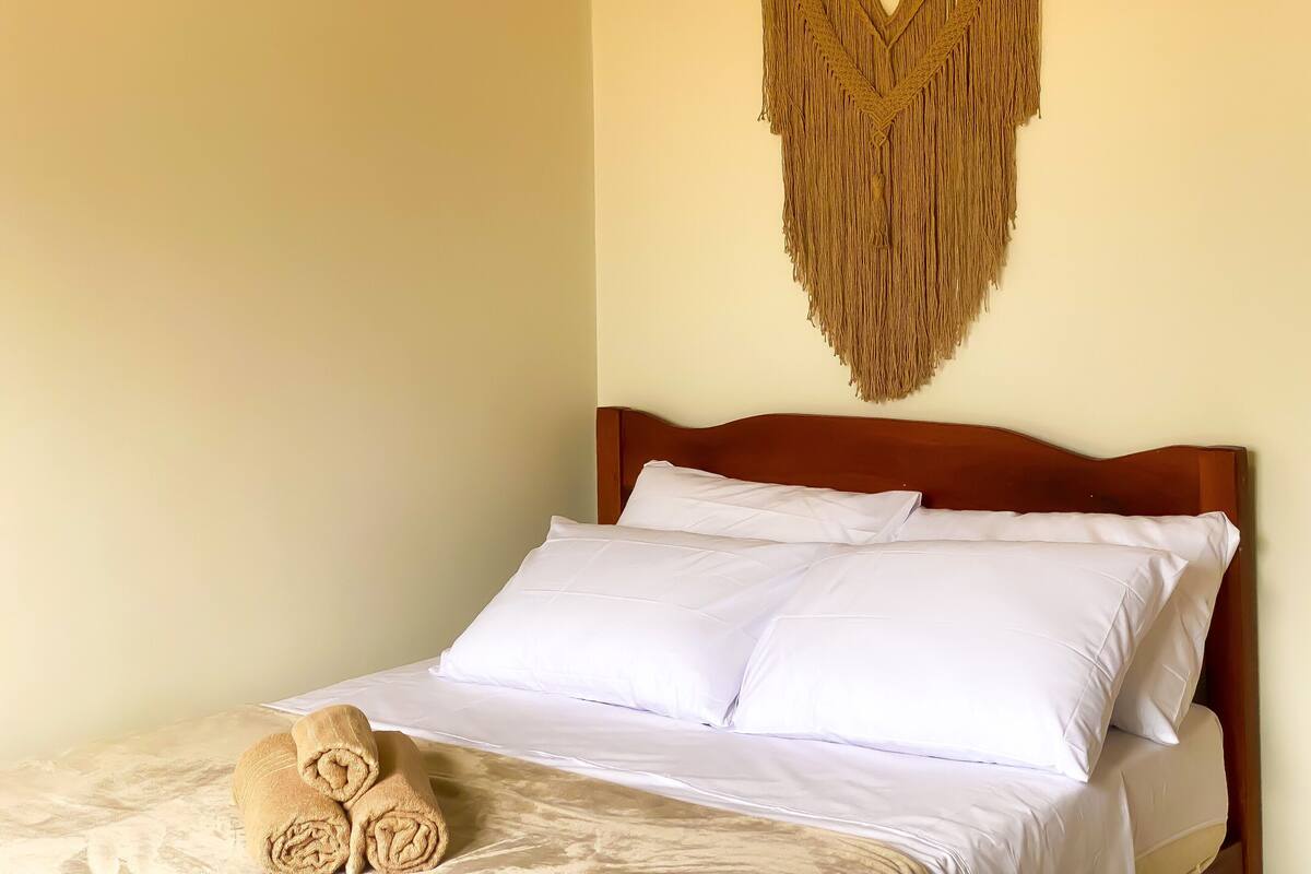 Un lit avec du linge de lit blanc et une tête de lit en bois, une décoration murale en macramé et des serviettes enroulées posées sur le lit.