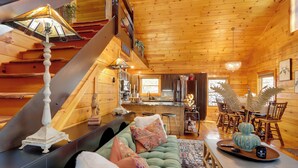 Interior acogedor de una cabaña de madera con un sofá verde, una mesa de comedor con sillas, una zona de cocina y una escalera.