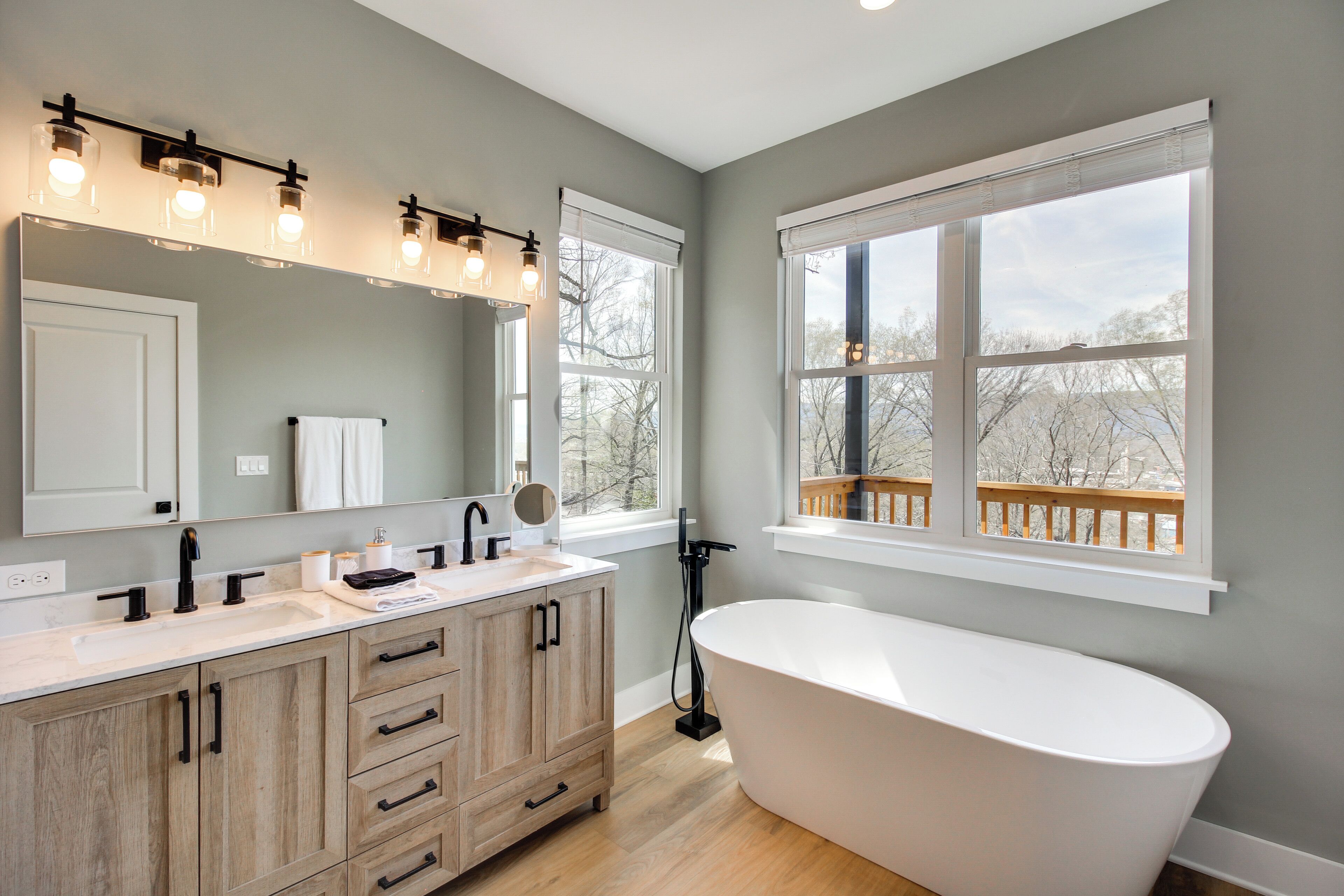 A modern bathroom with a freestanding bathtub, a vanity with a mirror and lights, and large windows offering a view of trees and a deck.