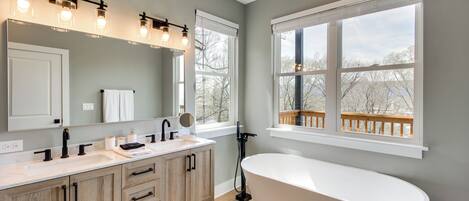 A modern bathroom with a freestanding bathtub, a vanity with a mirror and lights, and large windows offering a view of trees and a deck.