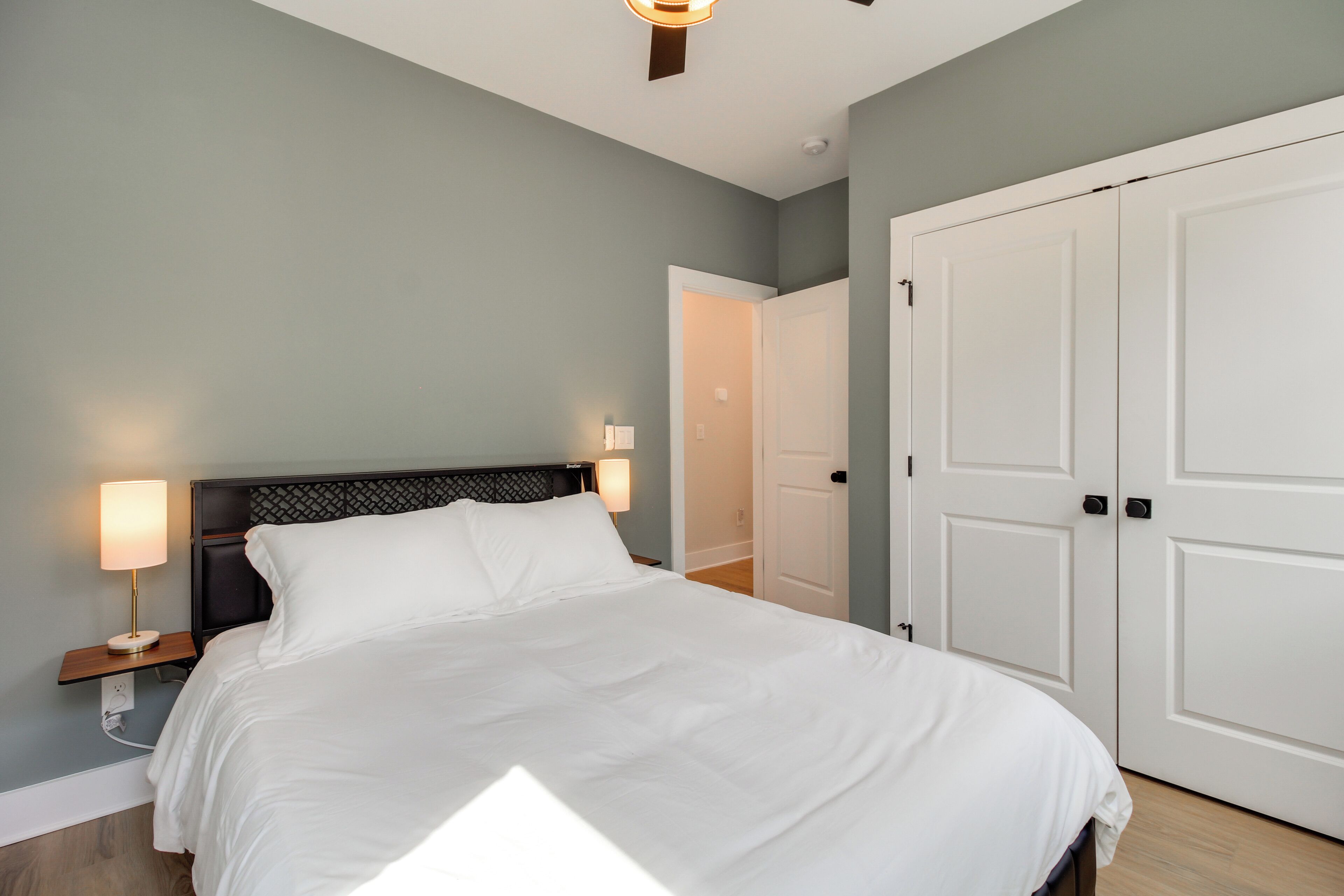 A neatly made bed with white linens, a nightstand with a lamp, and a closed door in a room with light blue walls.