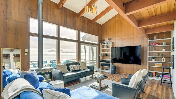 A spacious living room with a large window, a fireplace, a television, and a bookshelf. - Decks & Views: Waterfront Shelton Home (Shelton)