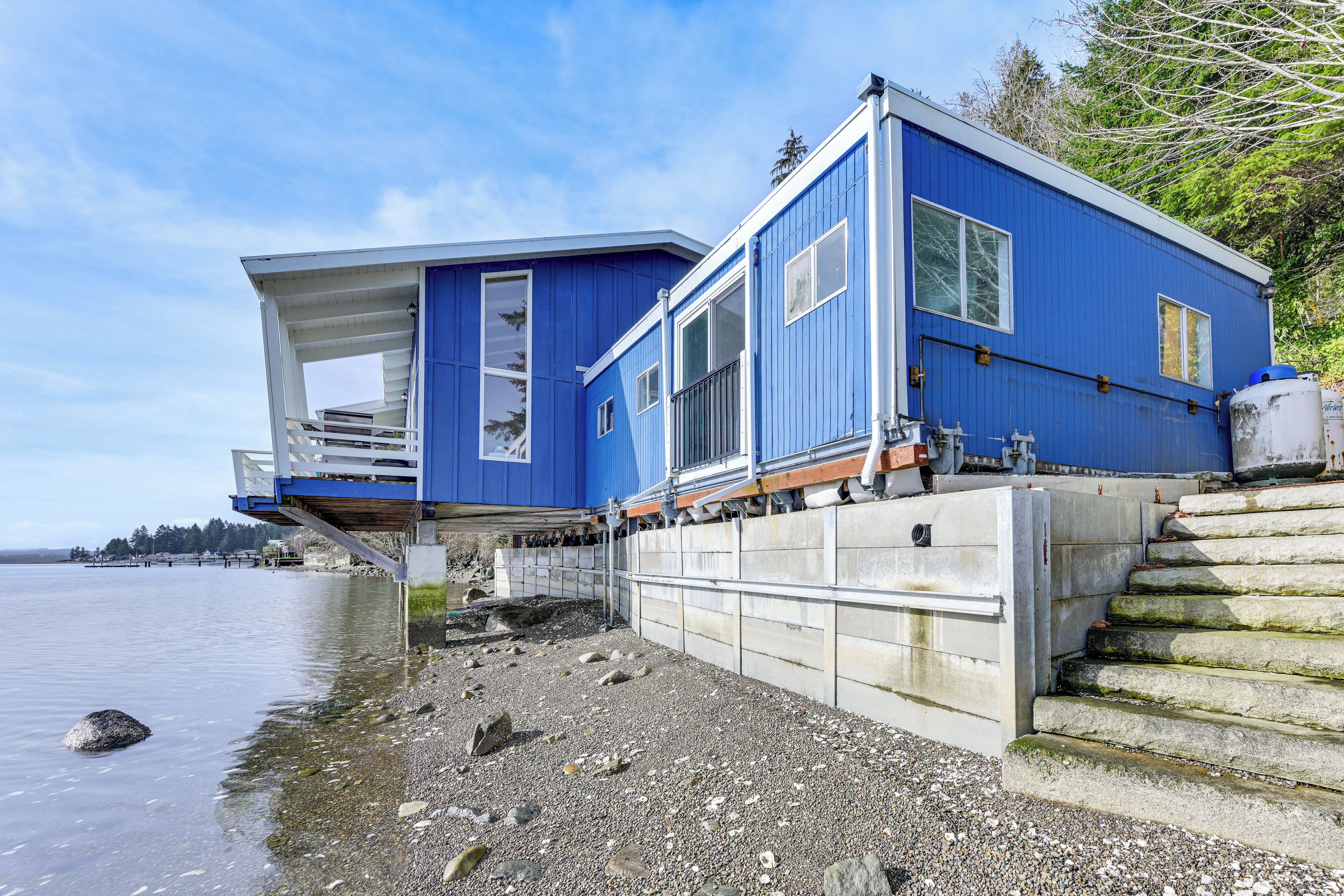 A blue waterfront house with a balcony, a concrete retaining wall, and a stone staircase leading to the entrance.