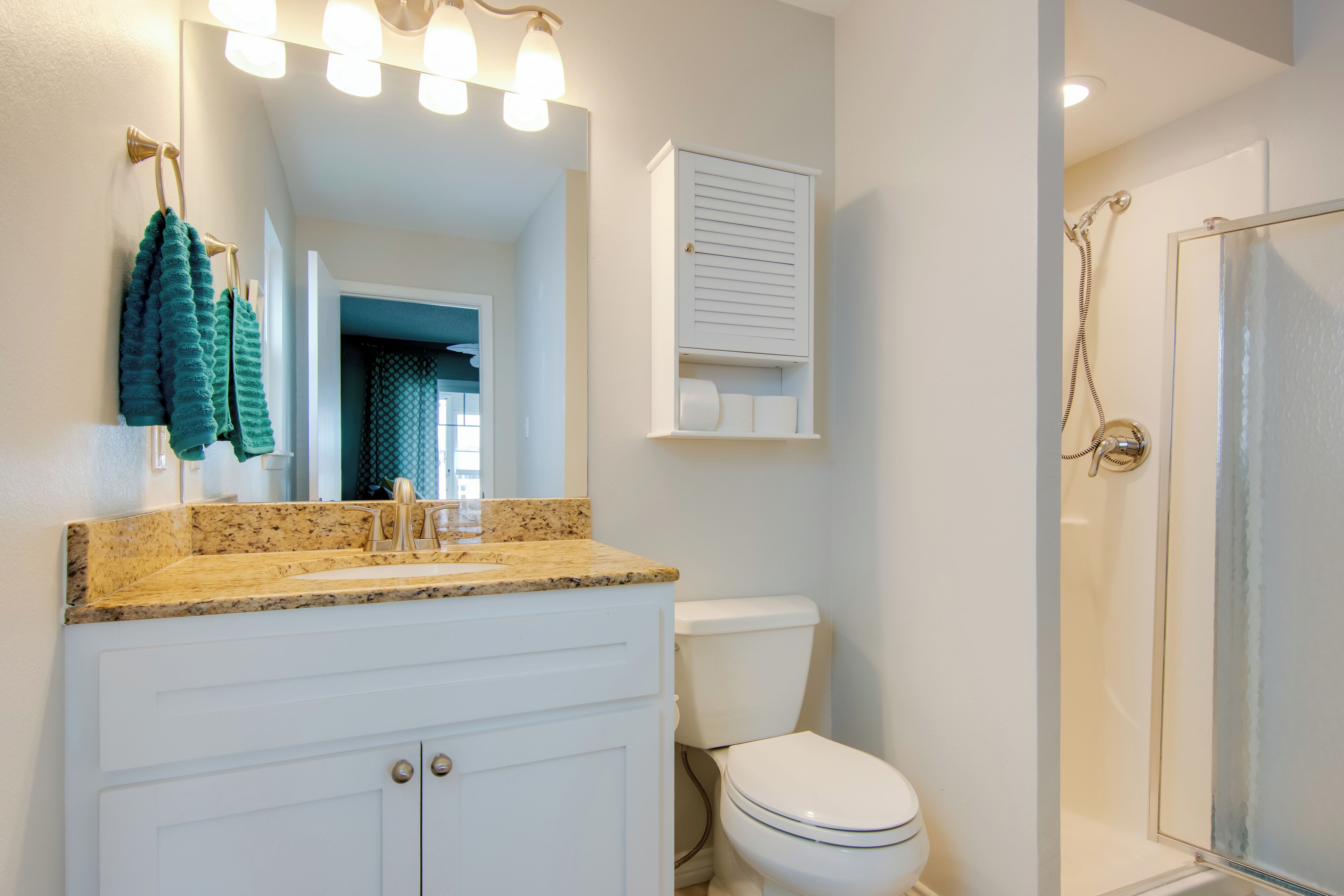 A bathroom with a granite countertop sink, white cabinets, a toilet, a glass shower enclosure, and a towel rack with green towels.