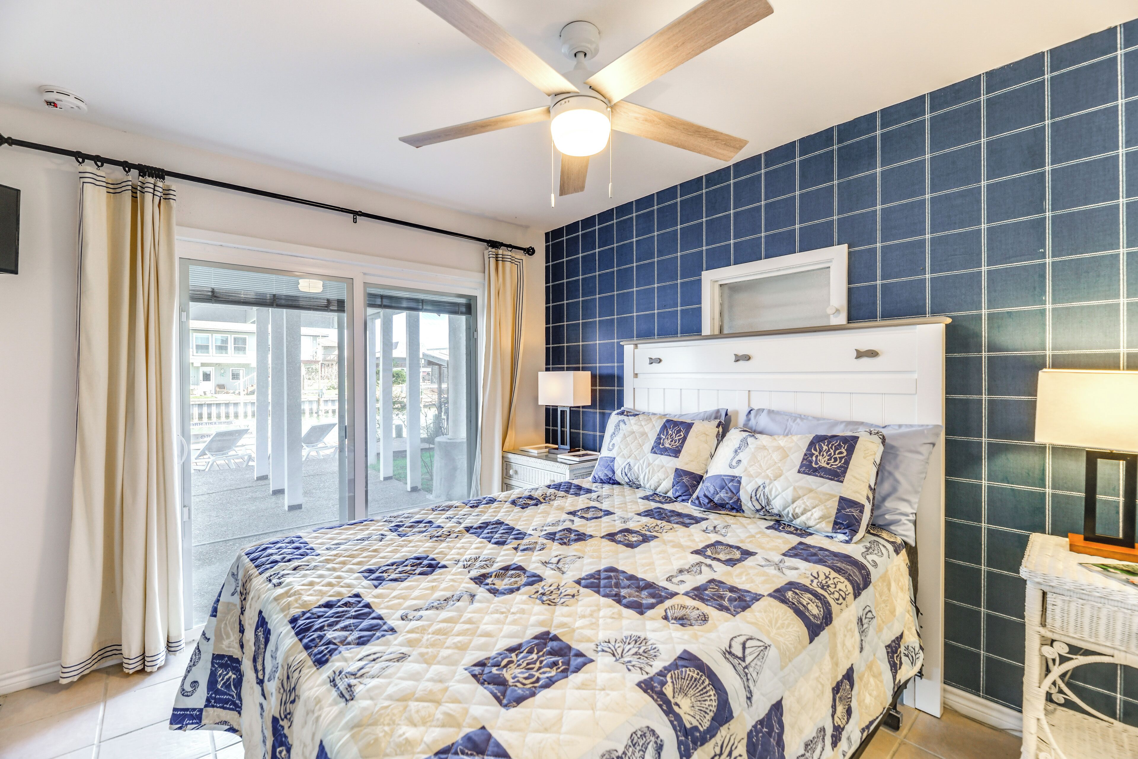 A bedroom with a large bed, a ceiling fan, a window with curtains, and a view of buildings outside.