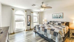 Une chambre avec un grand lit, un ventilateur de plafond, une fenêtre avec des rideaux et un tableau au mur.