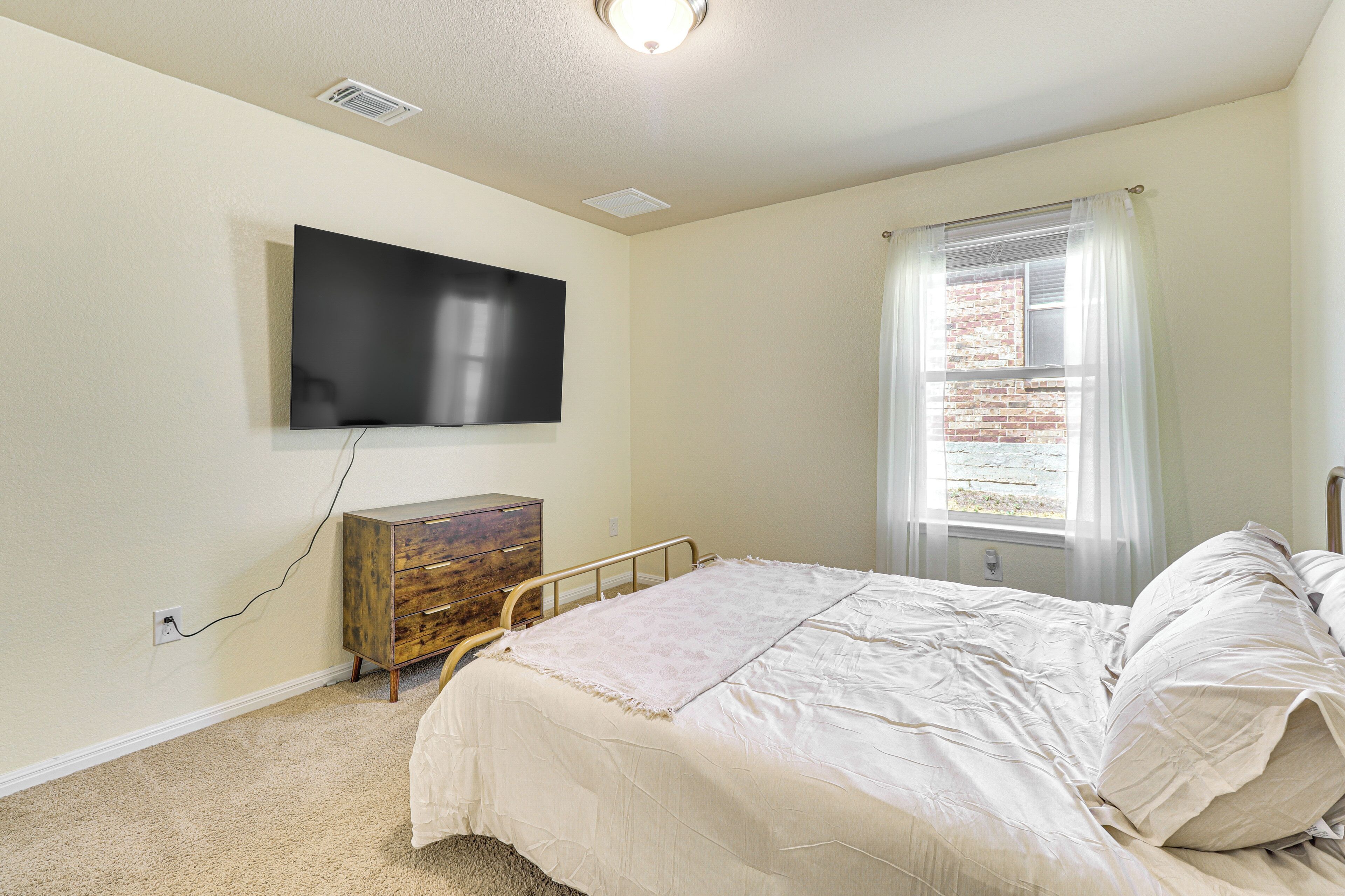 A bedroom with a bed, a dresser, a television mounted on the wall, and a window with curtains.