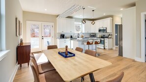 Una cocina moderna con armarios blancos, un comedor con mesa y sillas de madera, e iluminación colgante.