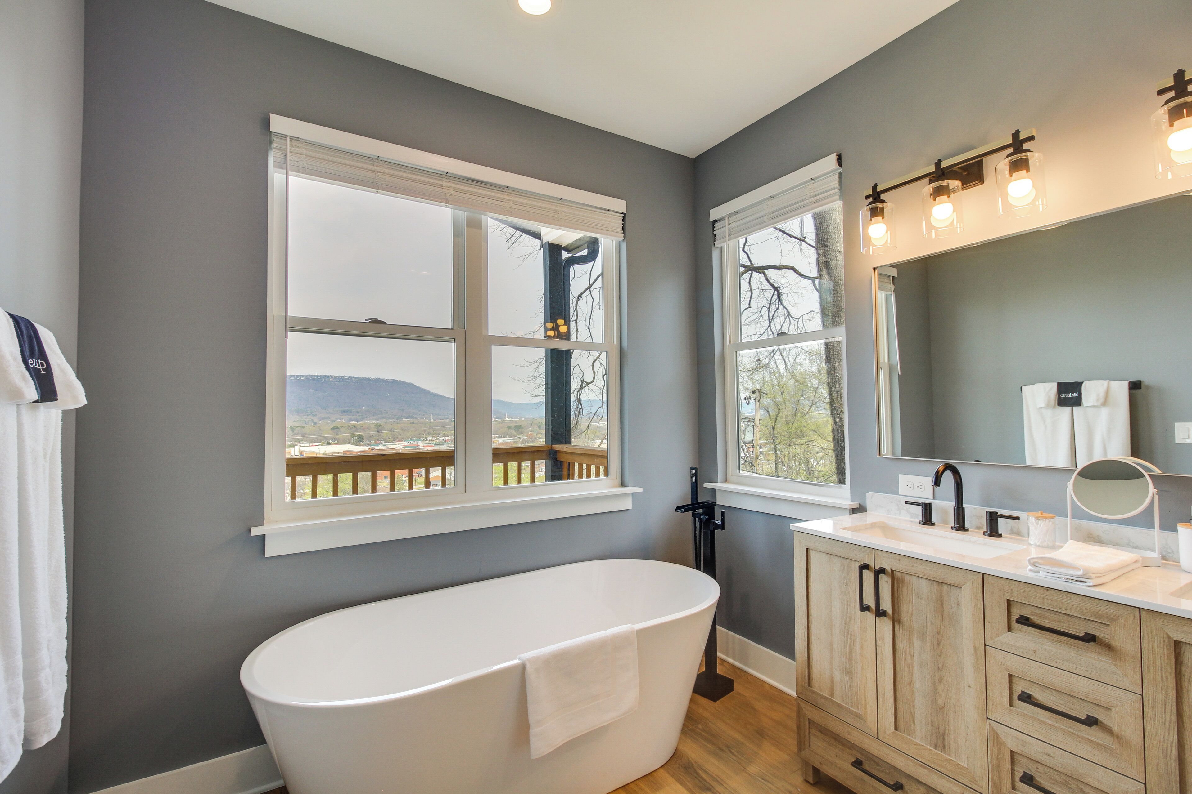 A bathroom with a freestanding bathtub, wooden cabinets, a vanity with a mirror, and large windows offering a view of the outdoors.