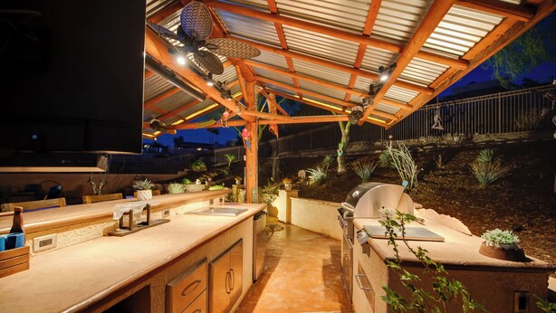 Uma área de cozinha externa com churrasqueira, banquetas de bar e uma estrutura de pérgula.