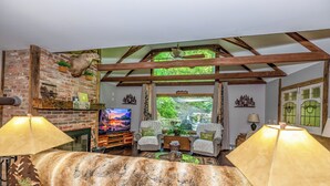 Una sala rústica con vigas de madera a la vista, pared de ladrillo, televisor, dos sillones, una manta de piel y vista a la vegetación exterior.