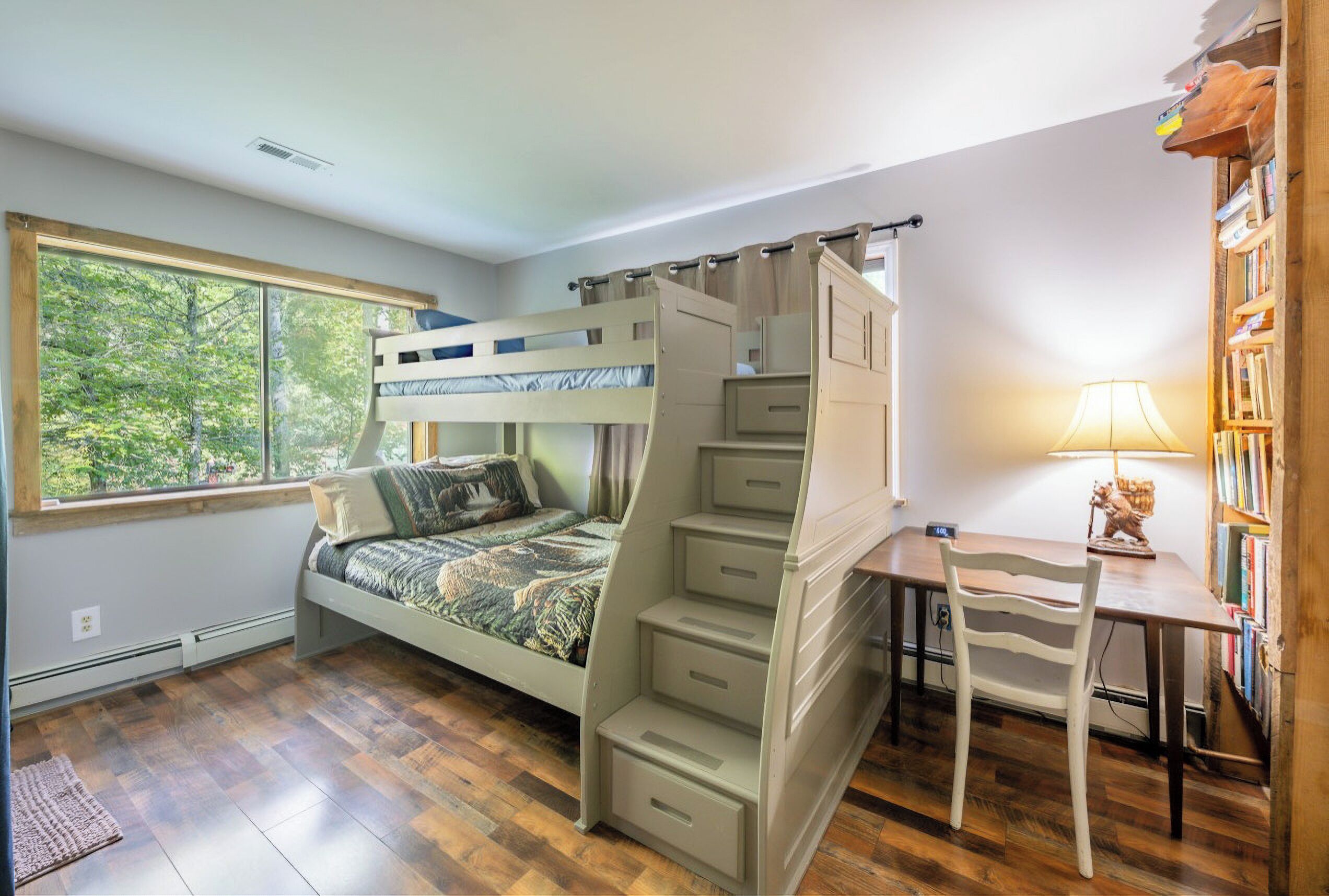 A bedroom with a bunk bed, a desk, and a chair. There is a window with a view of trees.