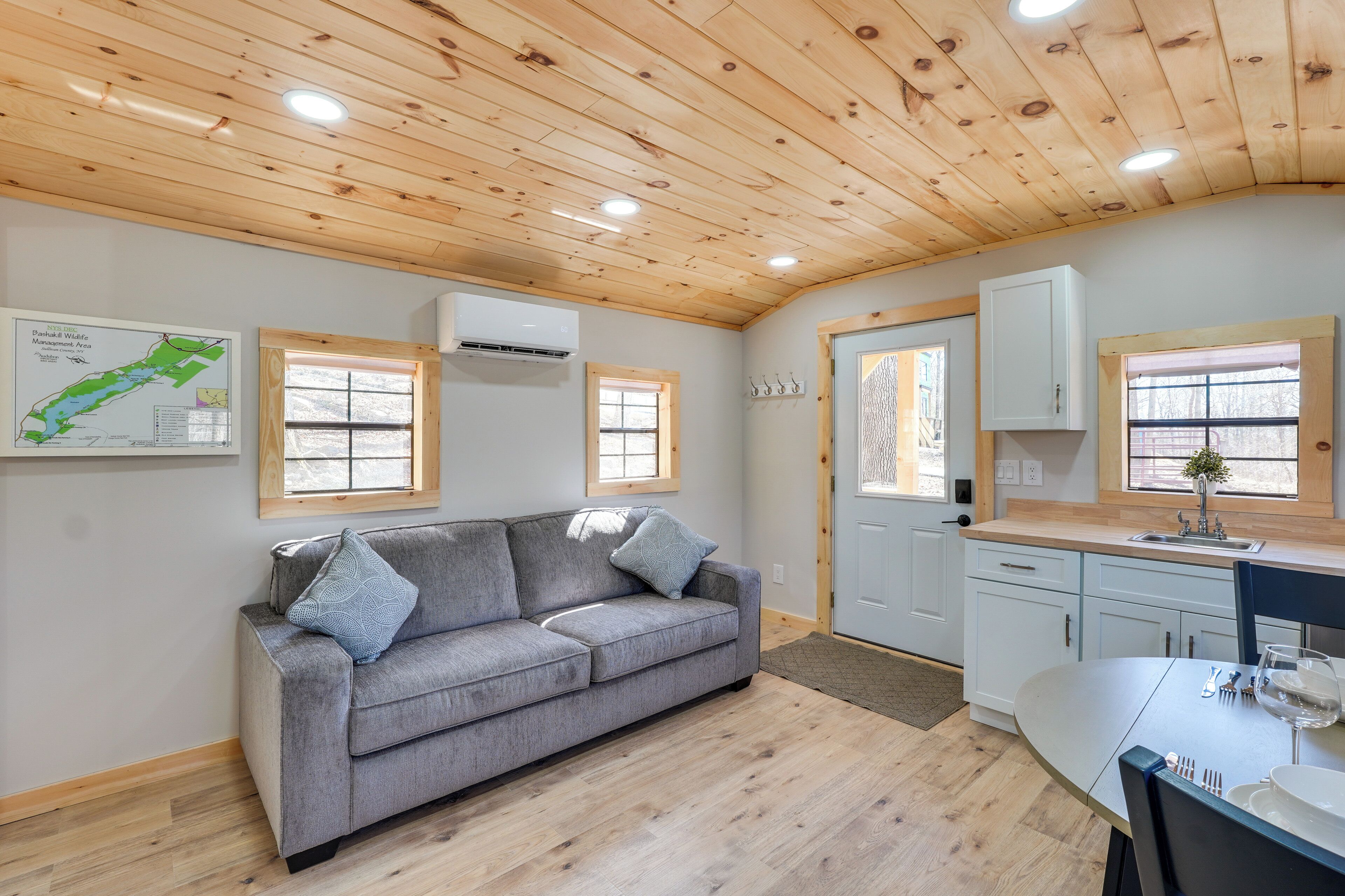 A cozy living room with a gray sofa, wooden ceiling, and a kitchen area with white cabinets and a sink.