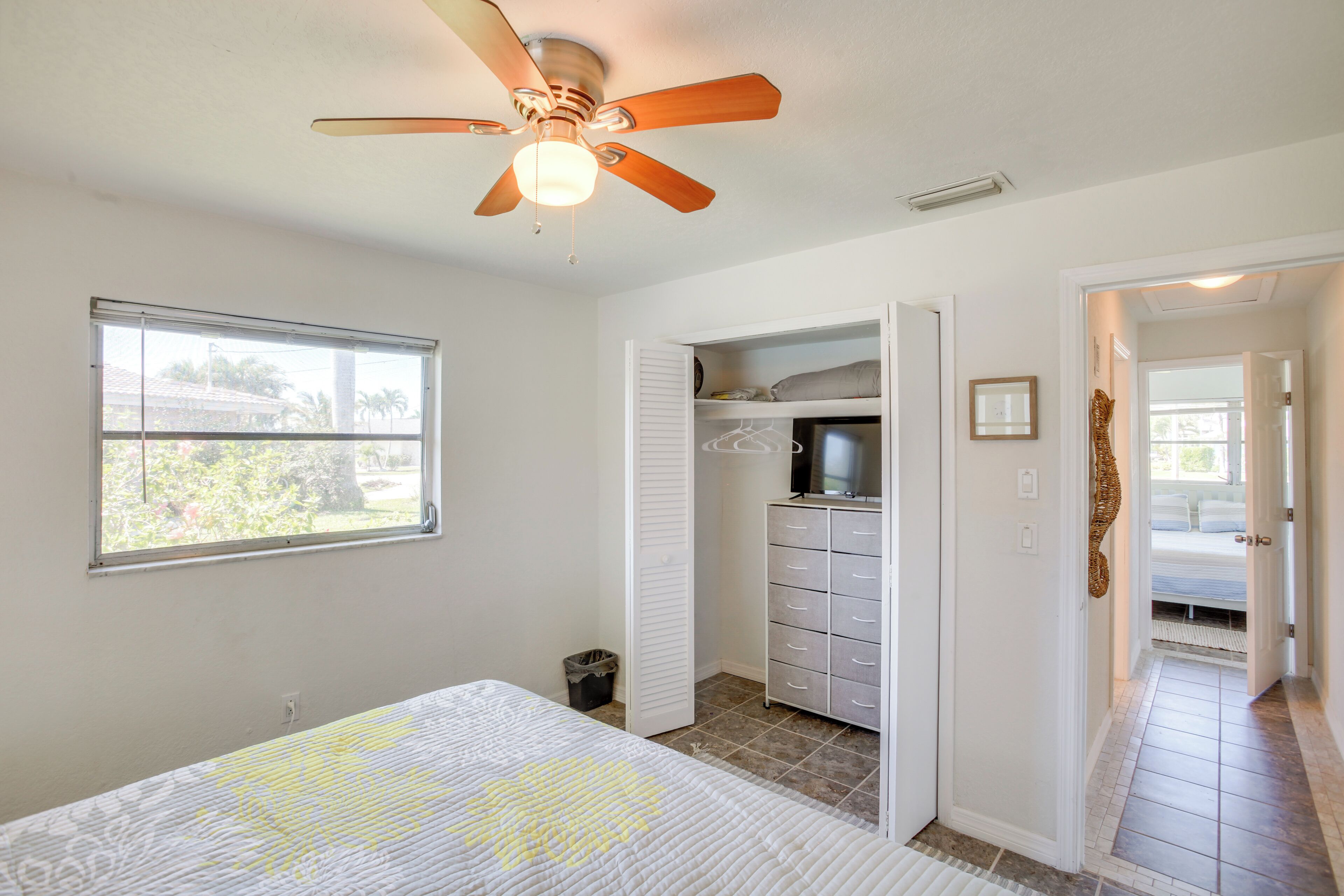 A bedroom with a bed, a ceiling fan, a window, a closet, and a hallway leading to another room.