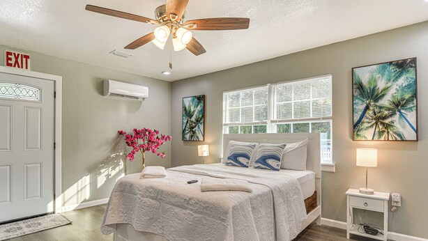 A bedroom with a bed, a nightstand, a ceiling fan, a window with blinds, and a door with an 'EXIT' sign. - Breezy Tampa Home w/ Yard: Perfect for Long Stays (Lutz)