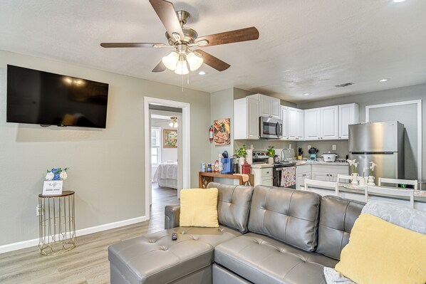 A modern living room with a grey sectional sofa, a ceiling fan, a flat-screen TV, and a kitchen area with white cabinets and stainless steel appliances.