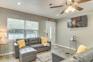 A living room with a grey sectional sofa, a small side table, a white rug, a ceiling fan, and a TV mounted on the wall.