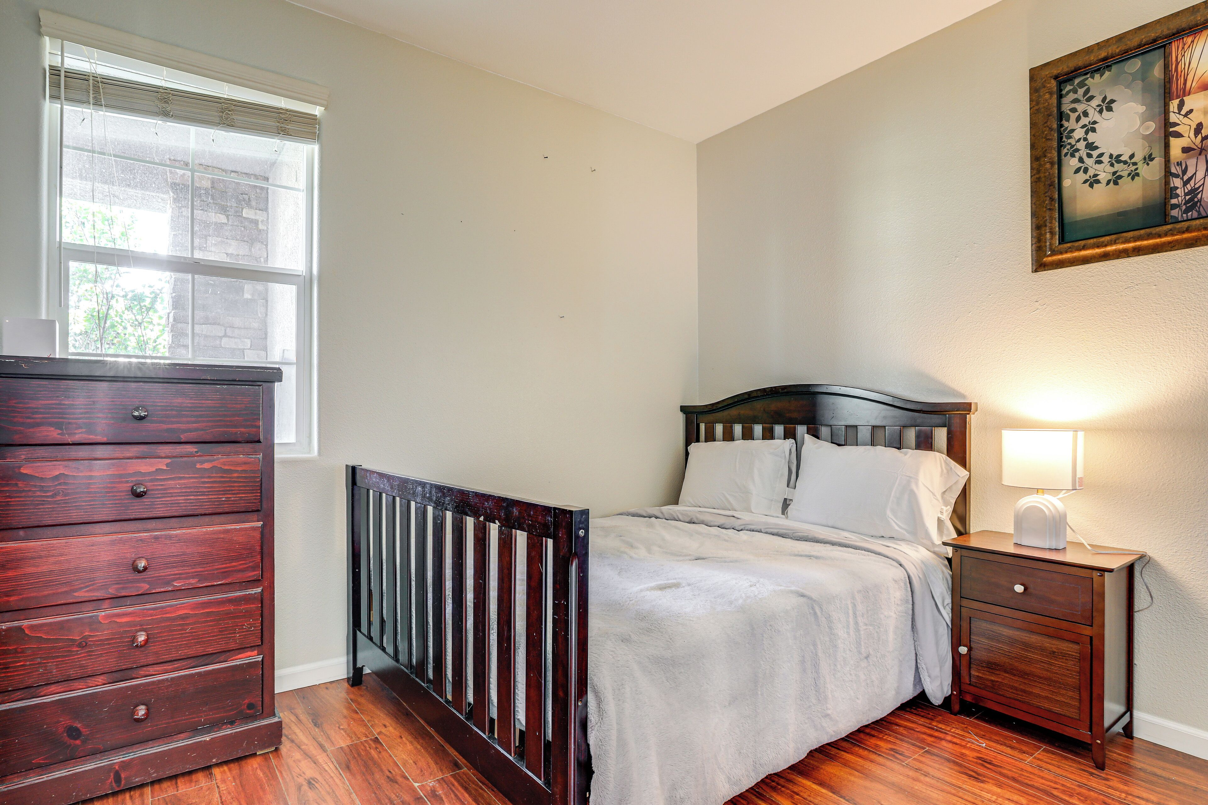 Une chambre à coucher avec une commode en bois, un lit avec une tête de lit, une table de chevet avec une lampe et une fenêtre avec des stores.