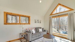 Un salon chaleureux avec un canapé gris, une petite table et deux fenêtres donnant sur une forêt.