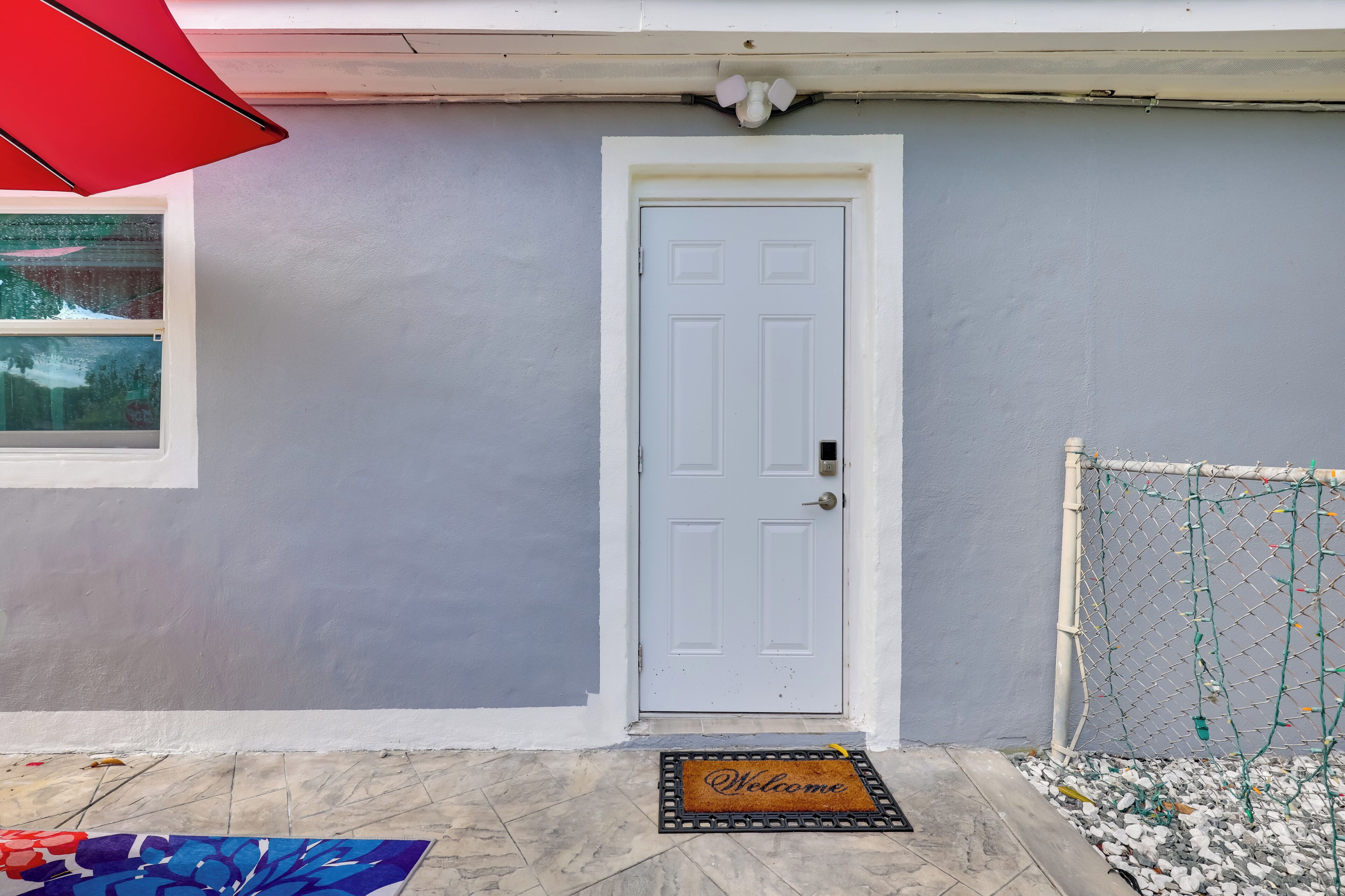 A white door with a doormat, a red umbrella, and a chain-link fence with greenery.