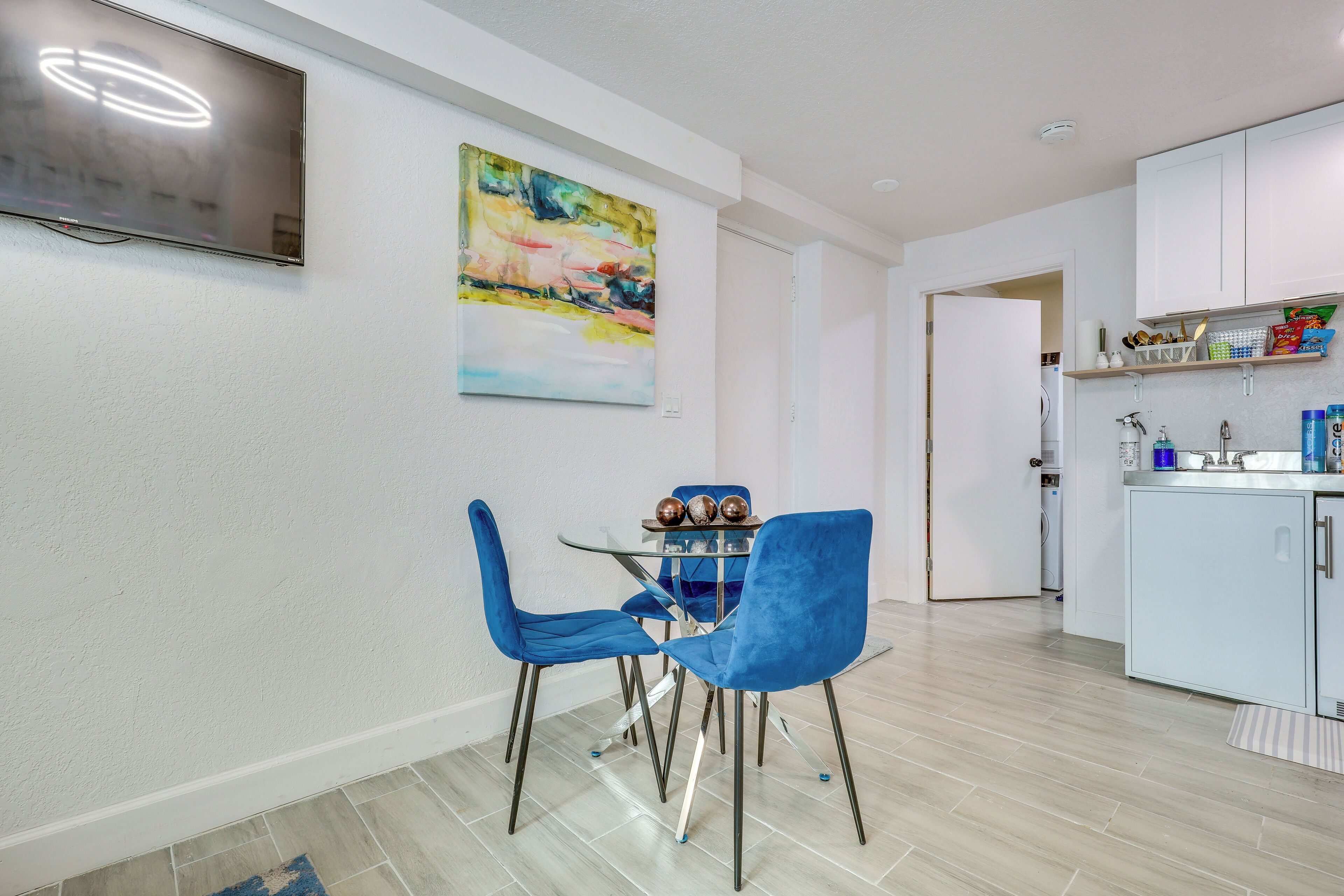 A modern kitchen with white cabinets, a dining area with blue chairs, and a flat-screen TV mounted on the wall.