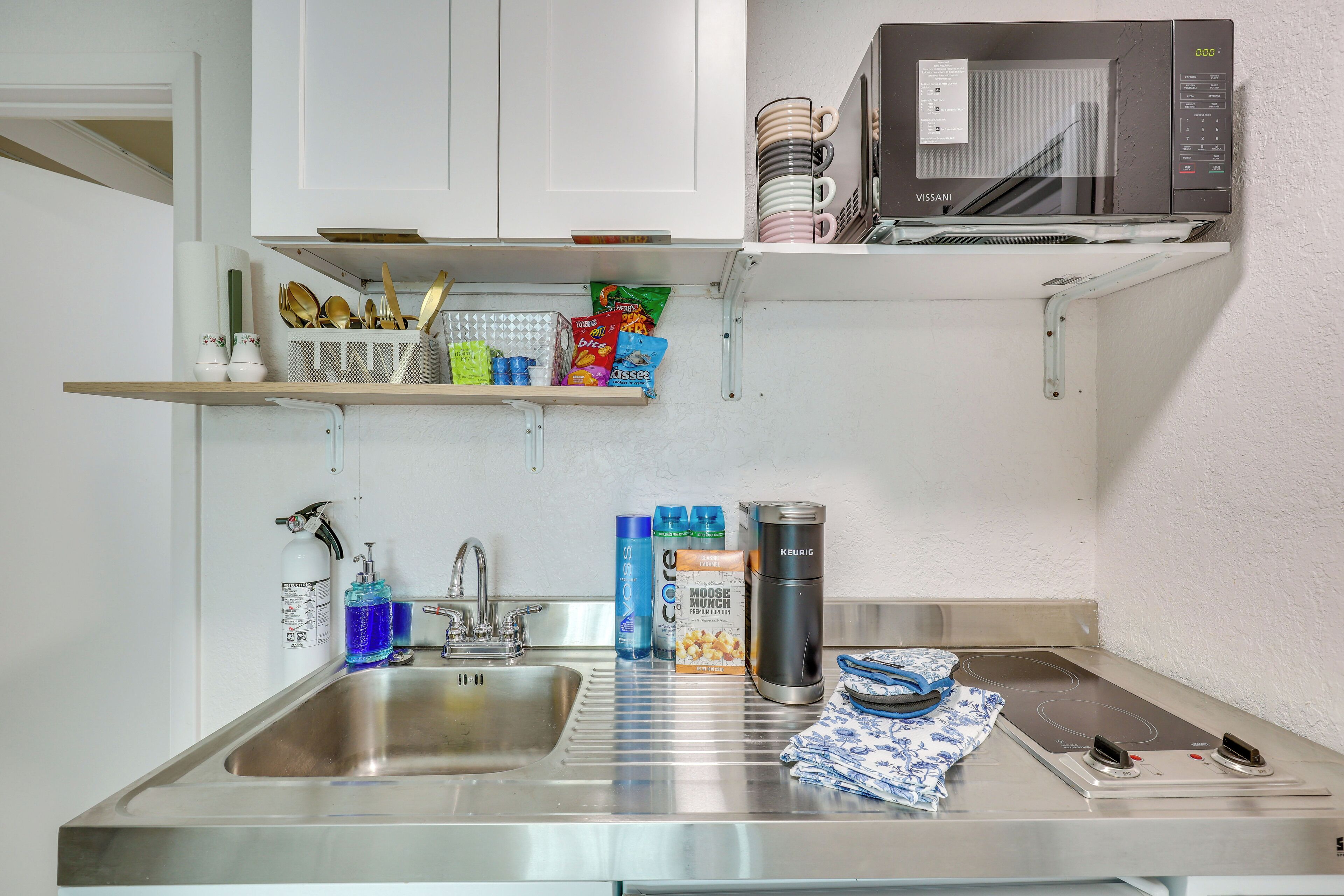 Una cocina compacta con un lavamanos de acero inoxidable, un microondas y un hervidor sobre la encimera.