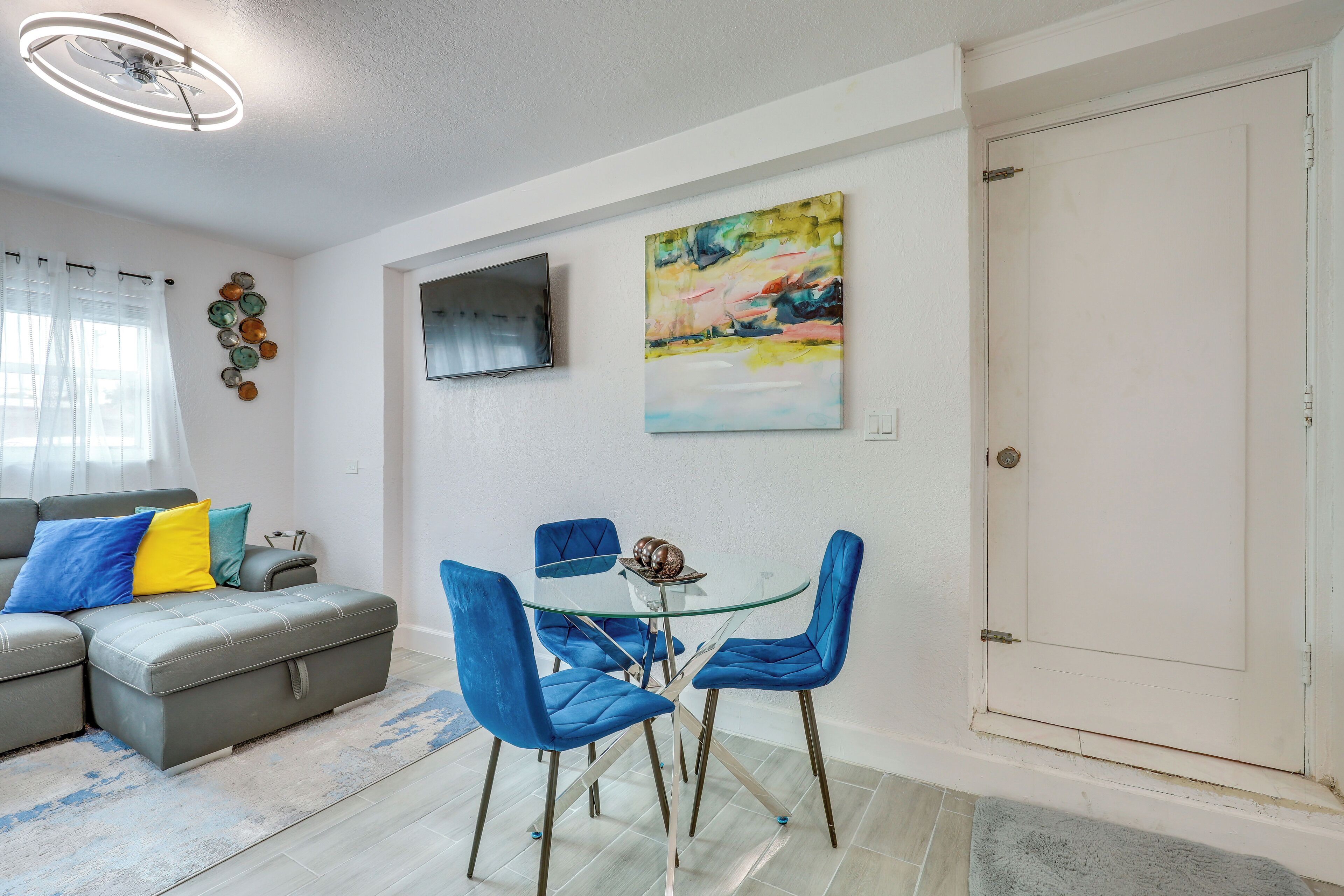 A modern living room with a grey sofa, blue chairs, a glass table, and a painting on the wall.
