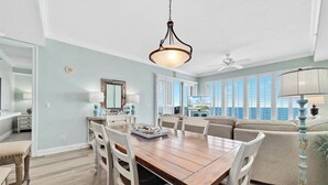 A spacious dining area with a wooden table and white chairs, a ceiling fan, and large windows offering a city view. - Regency Isle 1102 - Gulf Front Penthouse (Orange Beach)
