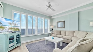 A spacious living room with a large window, a ceiling fan, and a beige sofa. - Regency Isle 1102 - Gulf Front Penthouse (Orange Beach)