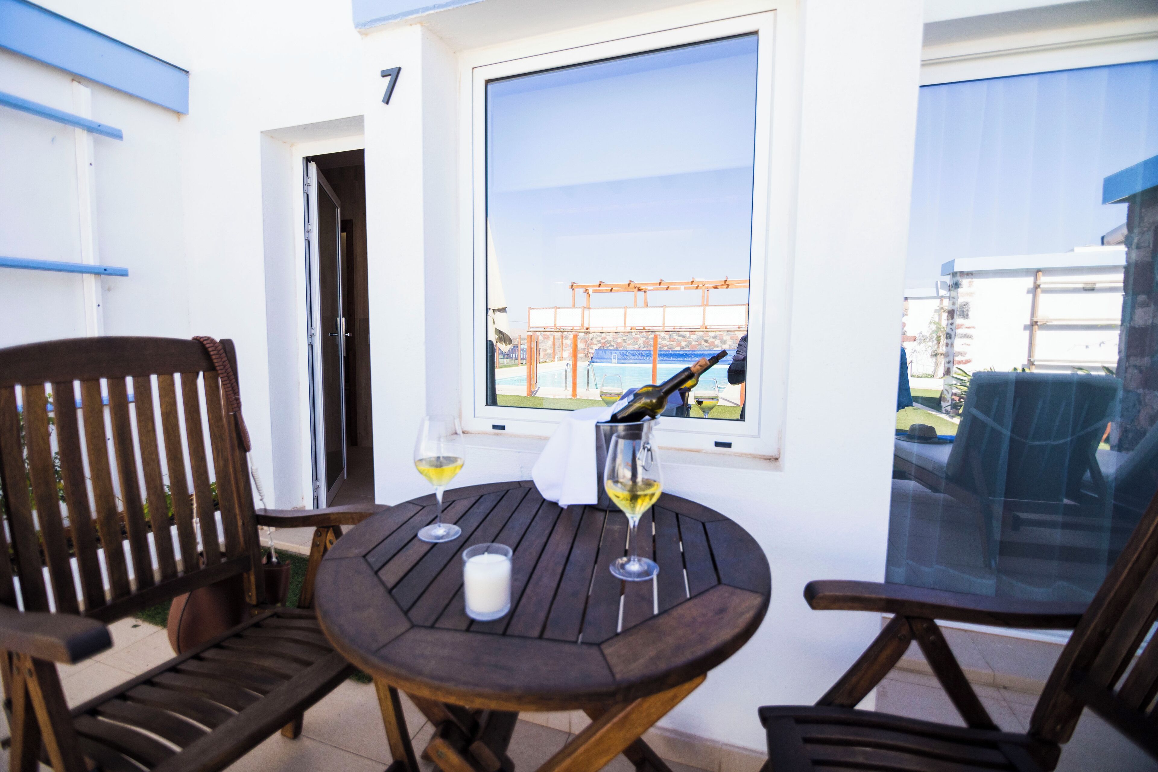 A wooden table with two wine glasses and a candle, a wooden chair, and a window with a view of a pool and buildings.