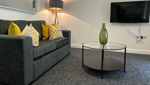 Un salon moderne avec un canapé gris, des coussins à motifs, une table basse en verre, un lampadaire et un téléviseur à écran plat fixé au mur.