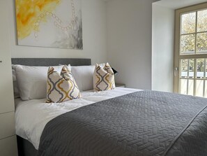 A bedroom with a bed, pillows, a quilted bedspread, a painting on the wall, and a window with a view.