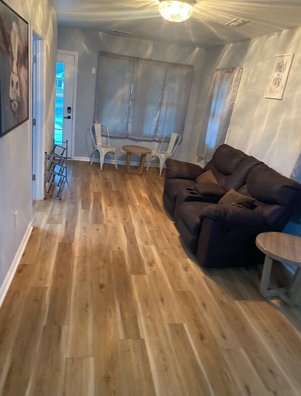 A living room with a brown sofa, a small round table, and a white chair. There is a wooden floor and a door leading to another room. - Charming 3-bedroom house in delightful Ravenel with WiFi, AC (Ravenel)