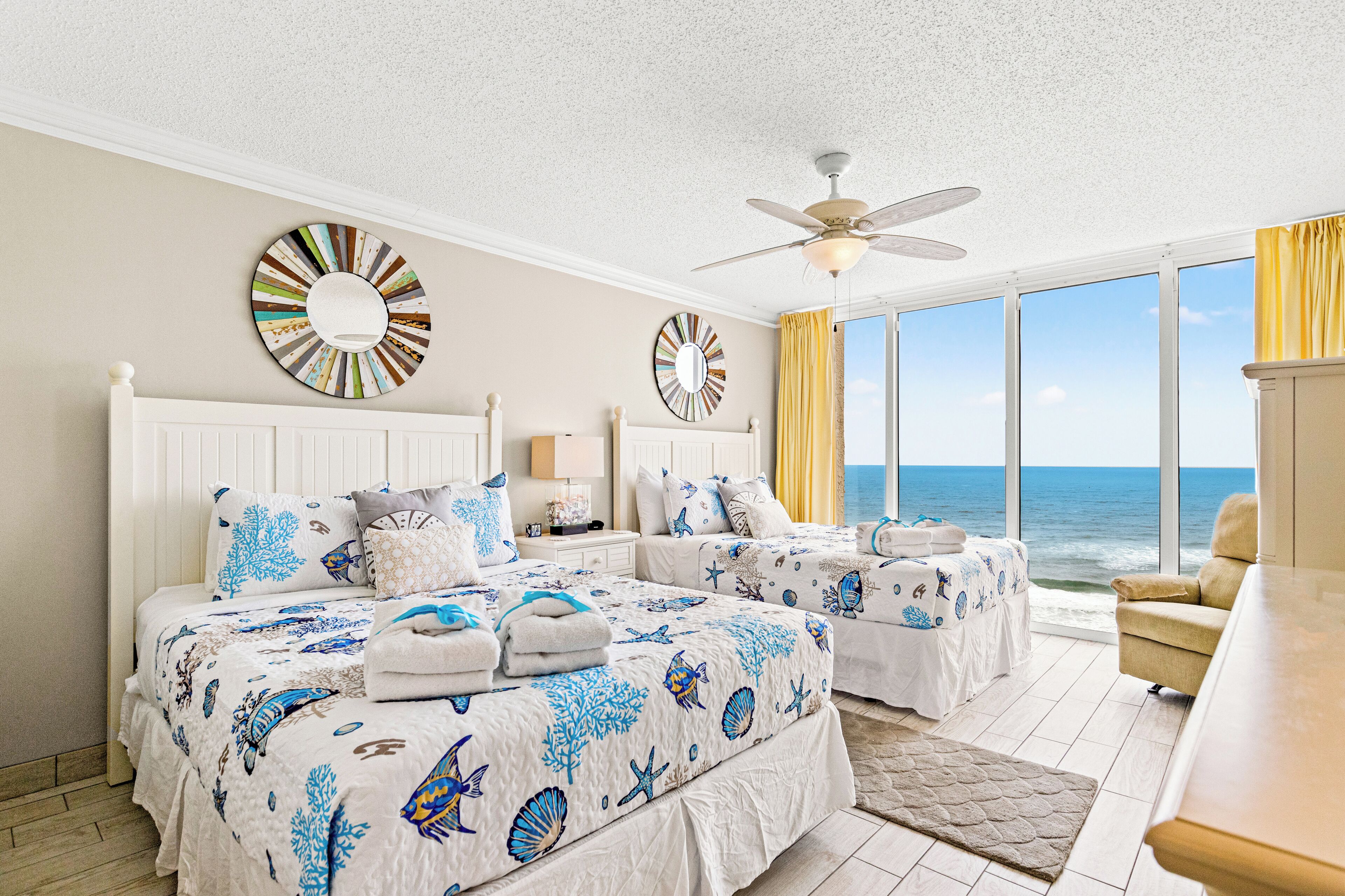 A bedroom with two beds, a ceiling fan, large windows with a view of the ocean, and decorative wall mirrors.