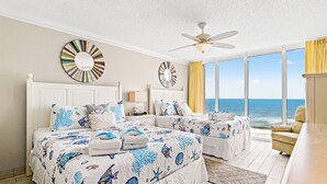 Un dormitorio con dos camas, ventilador de techo, ventanas amplias con vista al océano y espejos decorativos en la pared.