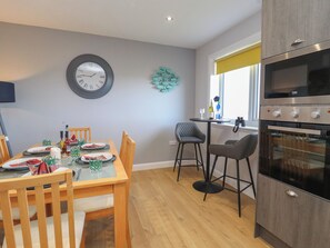 A modern kitchen with a dining area, wooden furniture, a clock, and a window with a yellow blind. - Lapwing Cottage (South Uist)