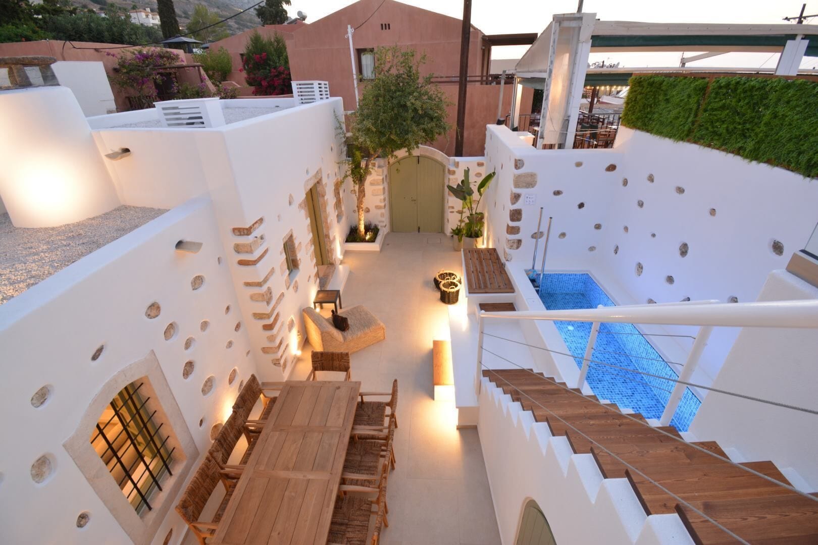 A modern outdoor pool area with a wooden deck and seating, surrounded by white walls with stone accents and a green hedge.