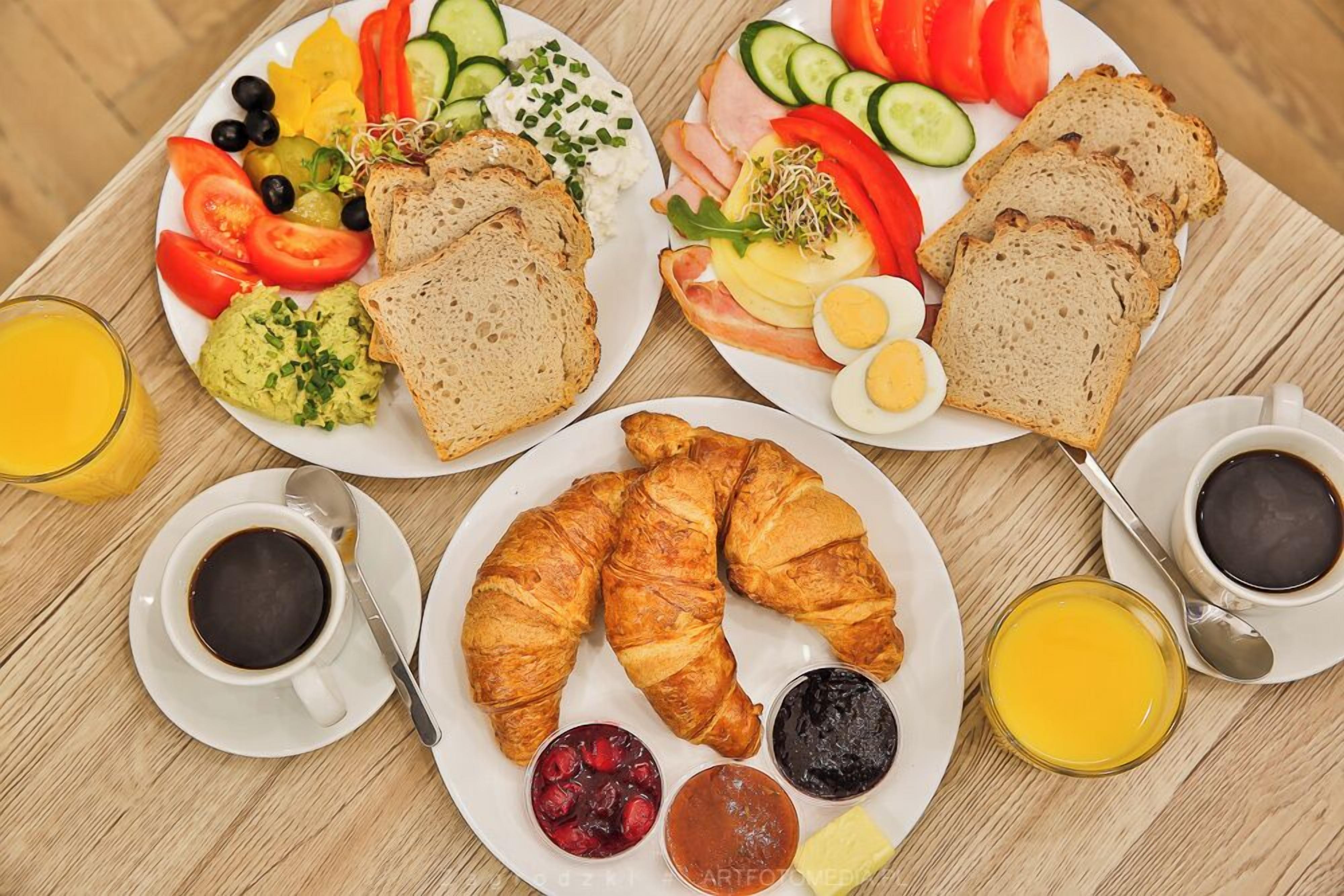 A breakfast setting with croissants, bread, eggs, and various fruits and vegetables on plates, along with cups of coffee and juice.