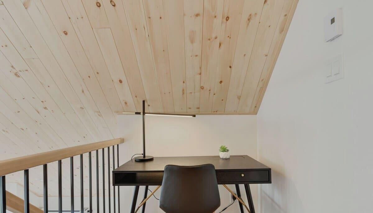 Una habitación con techo de madera, un escritorio con lámpara, una silla y una planta pequeña.