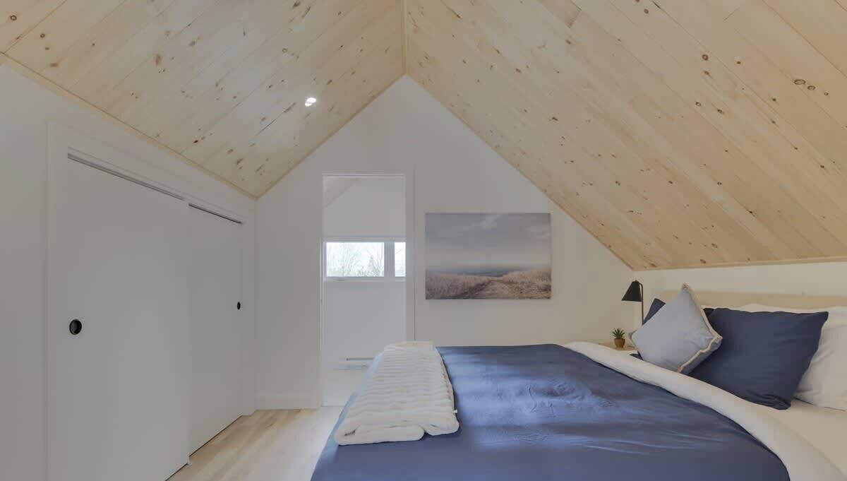Une chambre avec un plafond en pente, un lit avec une literie bleue, un plaid blanc et un tableau accroché au mur.