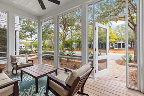 A covered outdoor area with a wooden deck, seating, and large windows offering views of a landscaped yard and neighboring properties.