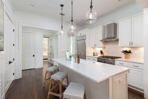 Una cocina moderna con armarios blancos, una isla grande con taburetes, iluminación colgante y electrodomésticos de acero inoxidable.