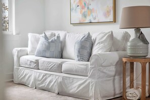 Um sofá branco com almofadas estampadas, um candeeiro sobre uma mesa de apoio de madeira e um quadro na parede.