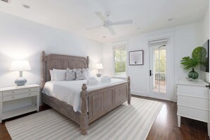 Quarto com cama de madeira, mesas de cabeceira brancas, ventilador de teto, janela com vista e uma planta verde.