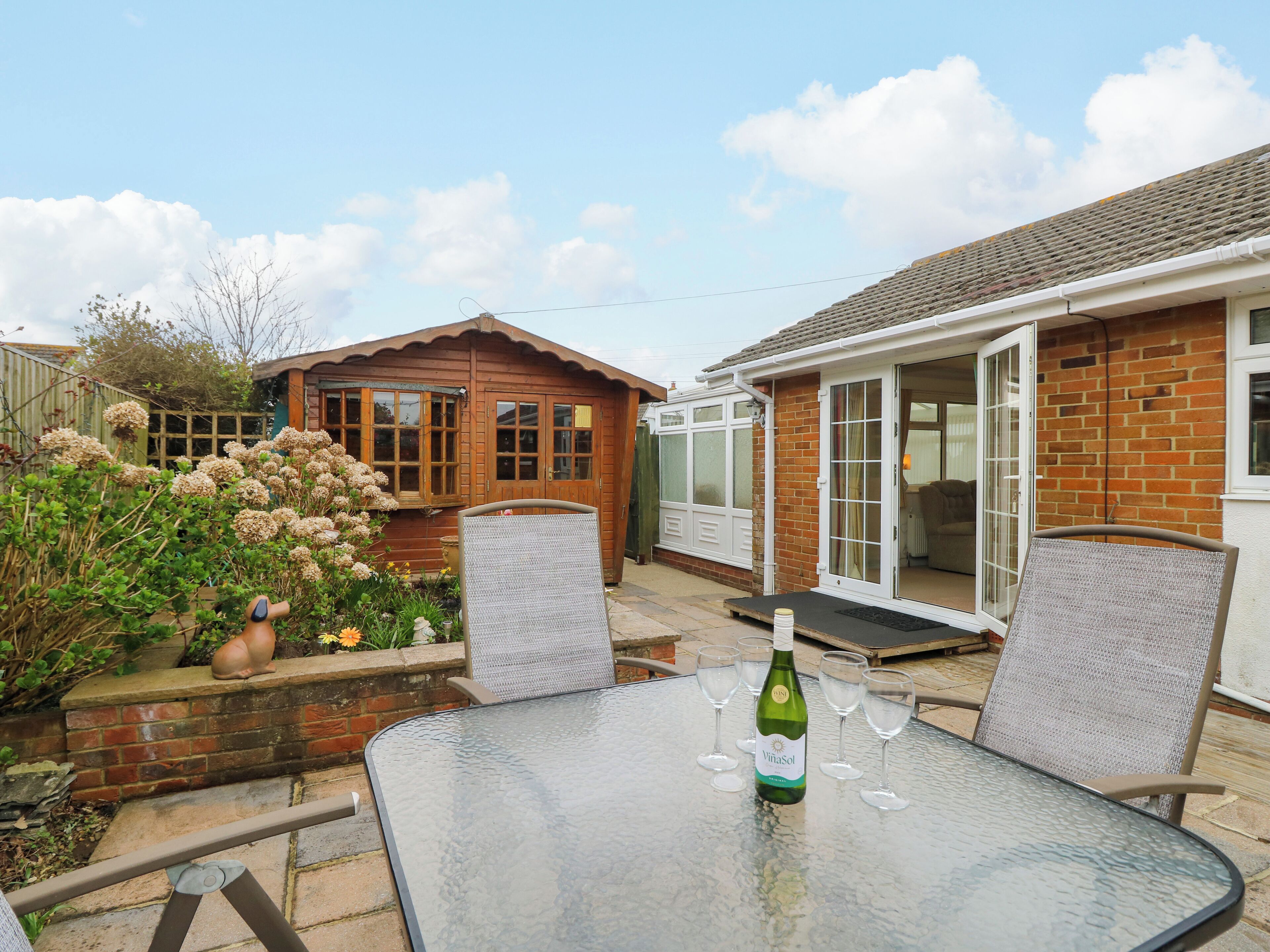 A glass table with a bottle of wine and glasses, a wooden house with a glass door, and a garden with flowers and a bird statue.