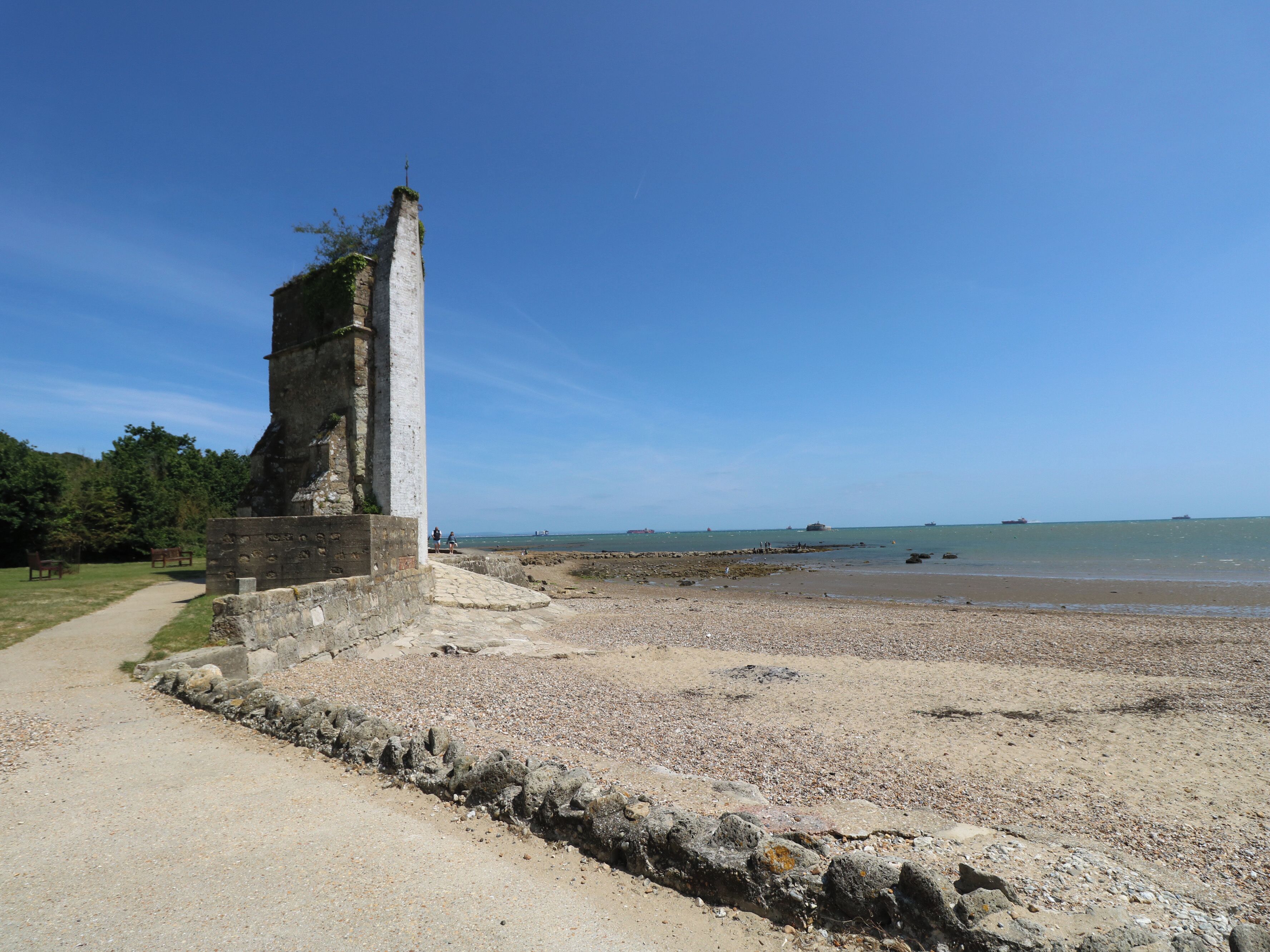 A coastal path leading to a stone monument by the sea, with a clear sky and distant ships.